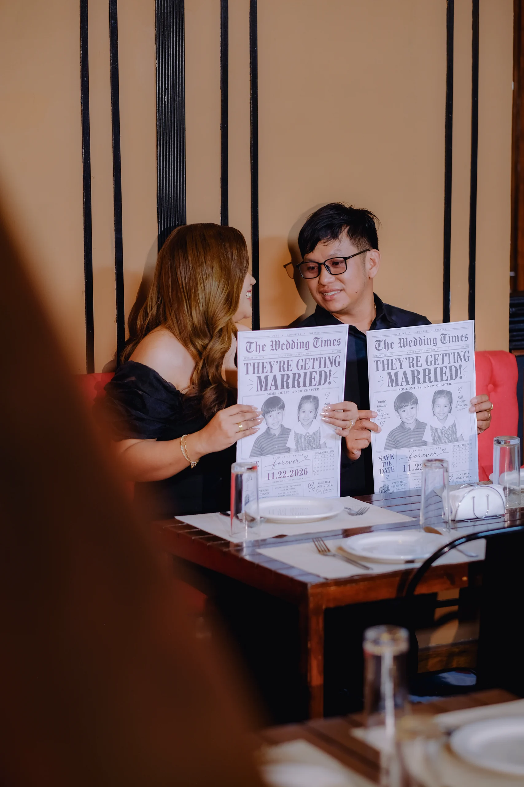 Ryan and Anne looking at each other with wedding newspapers on the red sofa.